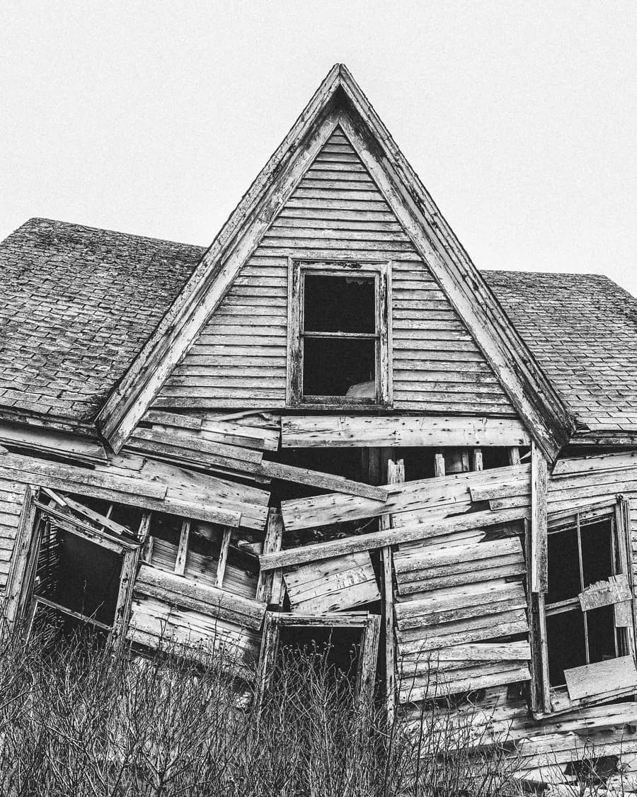 A collapsing wooden farmhouse, its face a patchwork of mismatched boards nailed over every opening, brush grown up to the porch line.