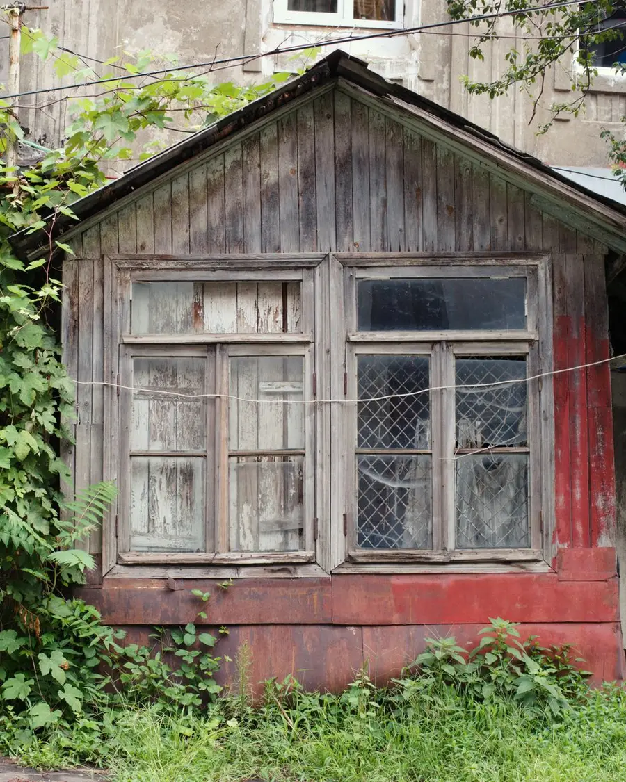 A small wooden cottage with peeling paint, vines taking the gable and weeds up to the window sills.